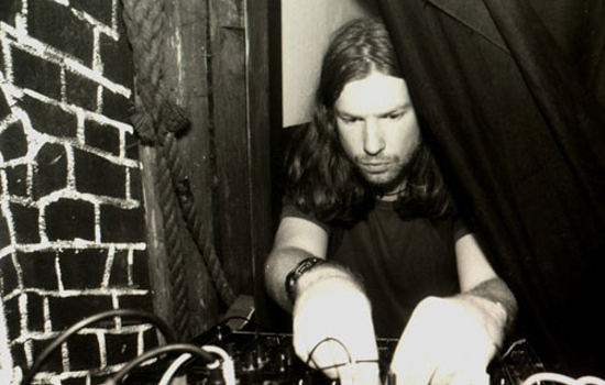 A monochrome photograph of a man with long hair and short facial hair playing a modular synth.